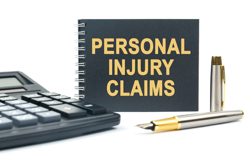 On a white background, there is a calculator, a pen, and a black notebook featuring the inscription "PERSONAL INJURY CLAIMS."