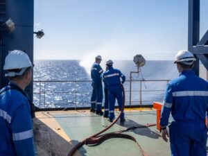 The image depicting seamen on board a merchant cargo ship during a fire emergency training drill, wearing firefighting equipment and handling a rigged fire hose.